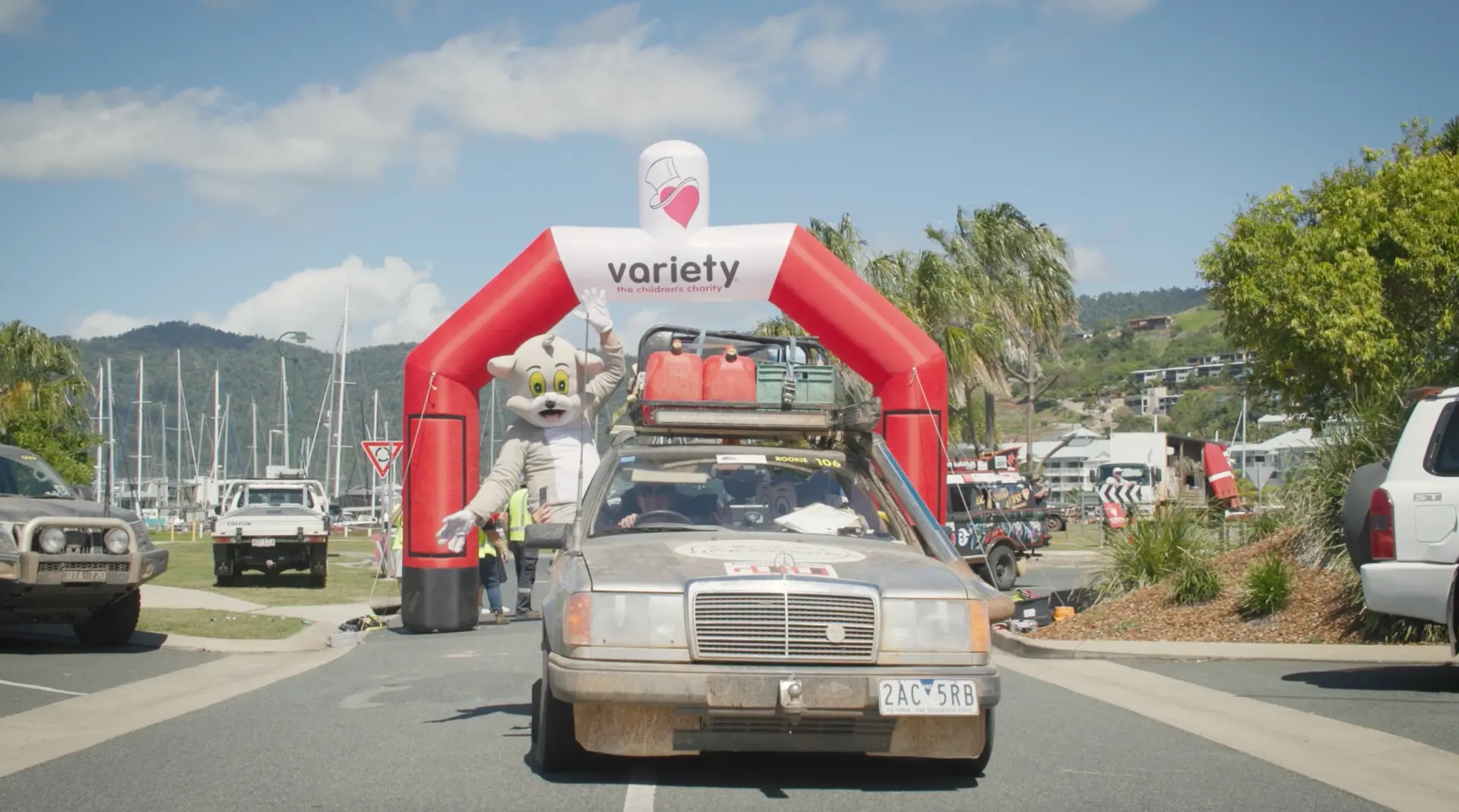Mud-splattered vintage Mercedes with a cat mascot waving from the window as it drives beneath a Variety inflatable arch at a coastal event start line