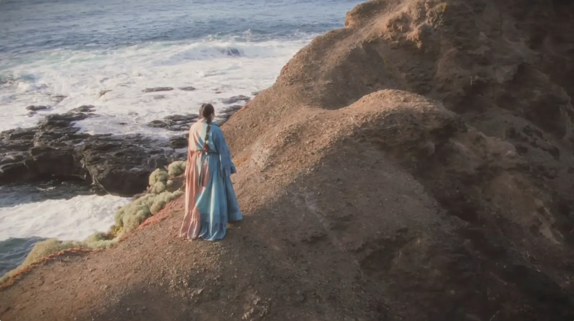 Aerial view of a figure in a flowing dress on a clifftop above the ocean